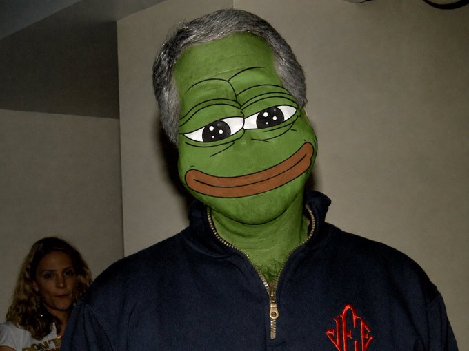 A man wearing a Pepe the Frog mask and a dark blue zip-up jacket stands indoors, with a woman partially visible in the background.