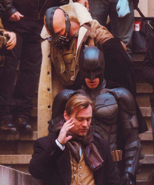 A behind-the-scenes shot shows actors in costume as Bane and Batman standing behind another man in a scarf and coat, with crew members visible in the background.