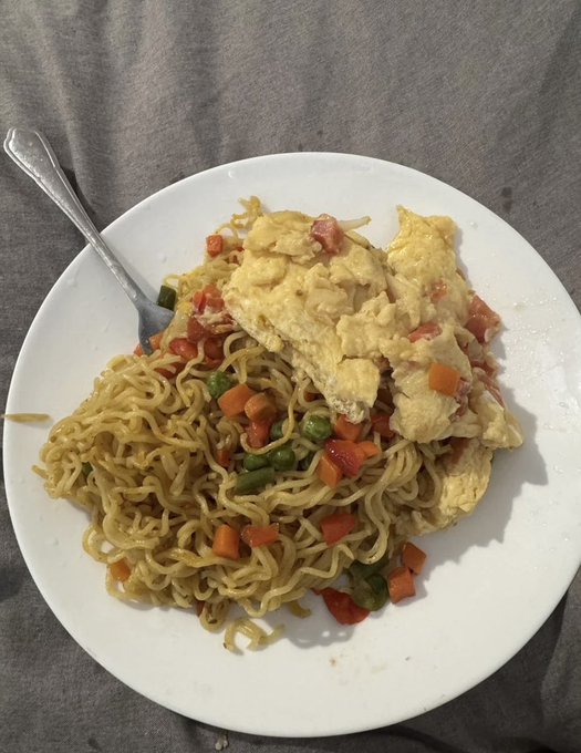 A plate of stir-fried noodles topped with scrambled eggs and mixed vegetables, including carrots, peas, and bell peppers, served with a fork on a gray surface.
