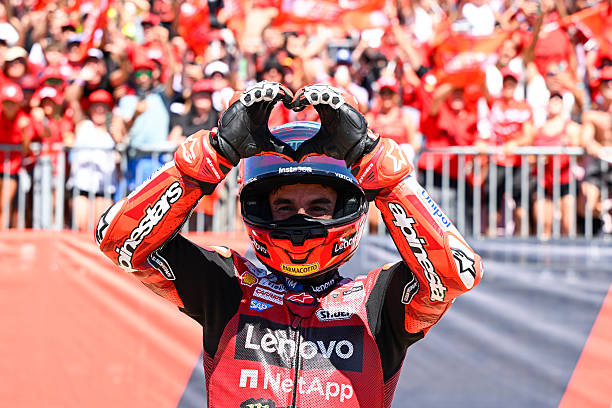 A MotoGP racer in a red Lenovo Ducati racing suit makes a heart shape with his gloved hands on his helmet, with fans in red visible in the background.