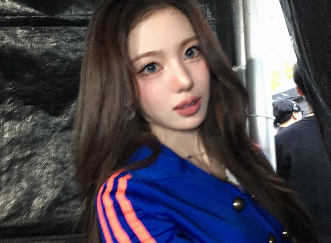 A selfie-style portrait of a young woman with long brown hair wearing a blue and orange striped jacket, with another person visible in the background.