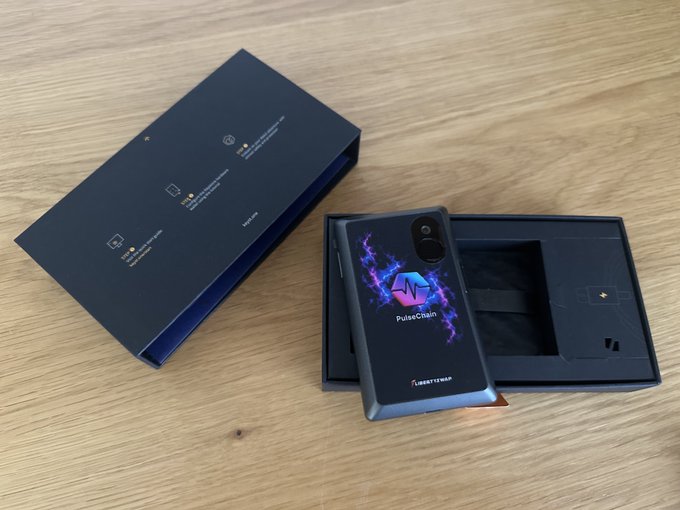A PulseChain-branded Keystone hardware wallet displays its vibrant purple and blue logo, freshly unboxed next to its sleek black packaging on a wooden surface—showcasing the self-custody experience mentioned in the announcement about upcoming firmware support and community feedback.