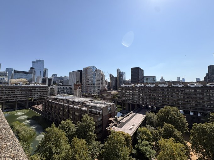 A wide daytime cityscape view featuring brutalist-style concrete apartment buildings in the foreground and a skyline of modern skyscrapers under a clear blue sky.