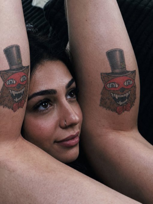 A close-up portrait of a woman with her arms raised, displaying matching tattoos on both forearms depicting a creepy, grinning red cat wearing a top hat.