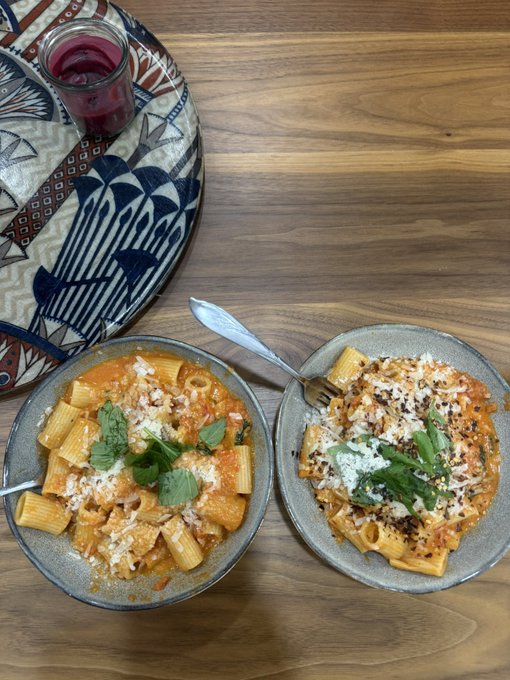 A top-down view of two plates of rigatoni in a creamy red sauce, garnished with grated cheese and fresh basil, served on a wooden table alongside a patterned cloth and a glass of red beverage. The hearty pasta meal makes for a comforting "Gm everyone" breakfast or brunch moment to start the day.