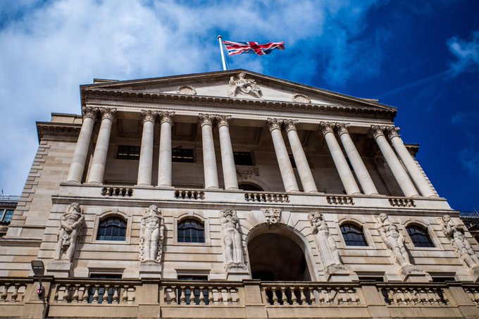 A grand neoclassical building with towering Corinthian columns, ornate sculptural figures, and the Union Jack flag flying prominently atop its pediment against a dramatic blue sky. The imposing architecture, characteristic of the Bank of England, provides a fitting backdrop to the news of the UK government's new directive empowering the central bank to support innovation in stablecoins and digital assets.