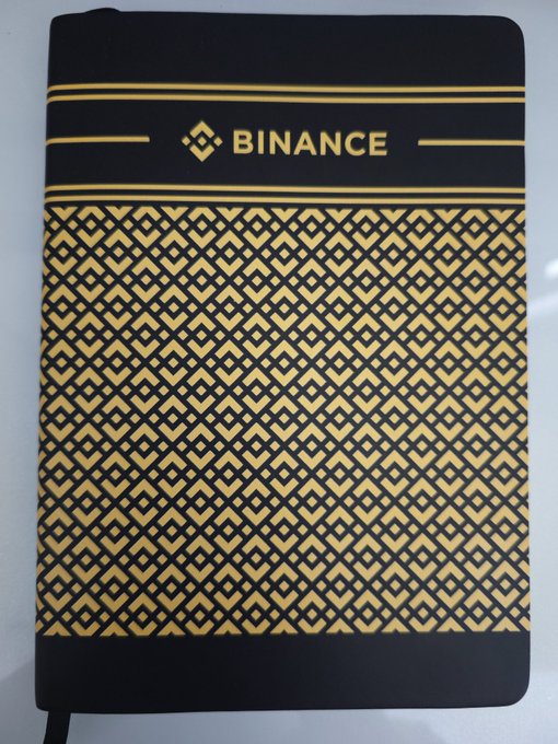 A black and gold Binance-branded notebook featuring the company's logo and diamond-pattern design, suggesting it's used to record daily cryptocurrency trading history as referenced in the post.