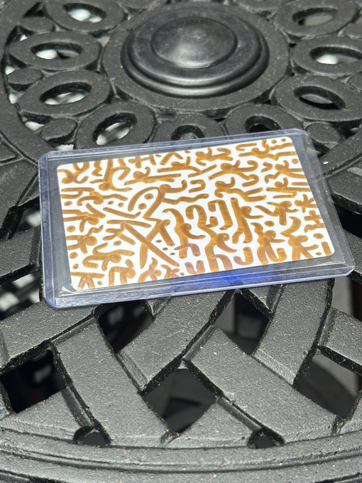 A decorative card with gold squiggle patterns on a white background sits in a clear plastic case on top of a black ornate metal surface.
