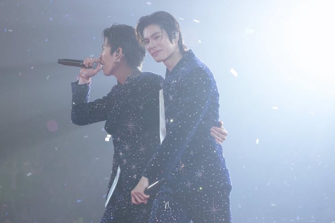 Two male performers in sparkly black outfits on stage with confetti falling, one holding a microphone while singing and the other embracing him from behind.
