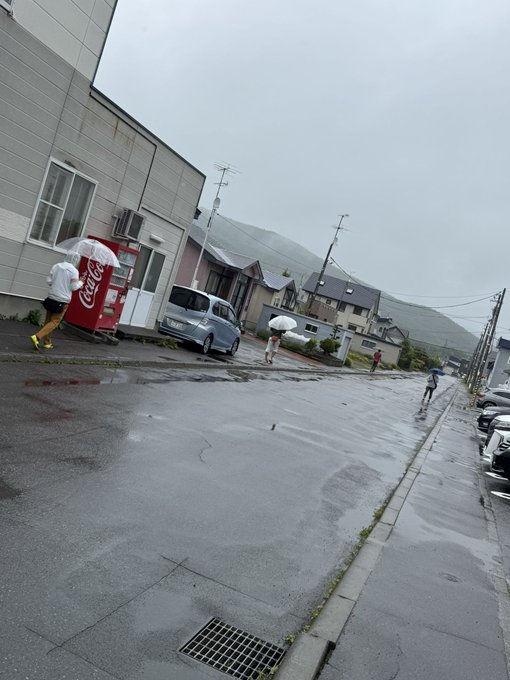 The image is tilted and shows a rainy street scene where a person in a white poncho tends to a red Coca-Cola vending machine on the left, while several runners in white jackets with umbrellas jog in formation down the wet road, fitting the post's description of runners in a line using STEPN energy in the rain the day before the Saroma Lake Ultra marathon.