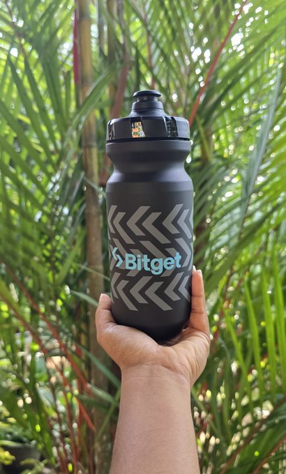 A hand holds a black Bitget water bottle with grey chevron patterns in front of green tropical foliage.
