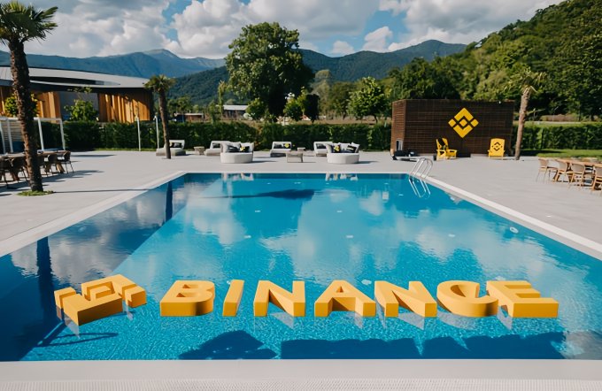 The image shows a resort-like setting with a pool featuring floating yellow letters that spell out "BINANCE". The post "GM七月你好" translates to "Good Morning July Hello" and suggests a greeting for the beginning of July, likely tied to an event or promotion by Binance during that month.