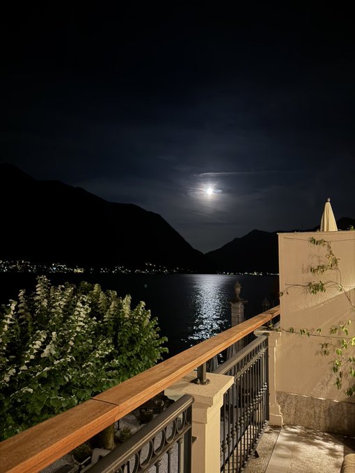 A moonlit lakeside view from a balcony terrace, with the moon’s reflection shimmering on the water beneath dark mountain silhouettes—mirroring the post’s message of tranquility and stillness, much like $BTC’s flat, low-conviction performance during the narrator’s trip.