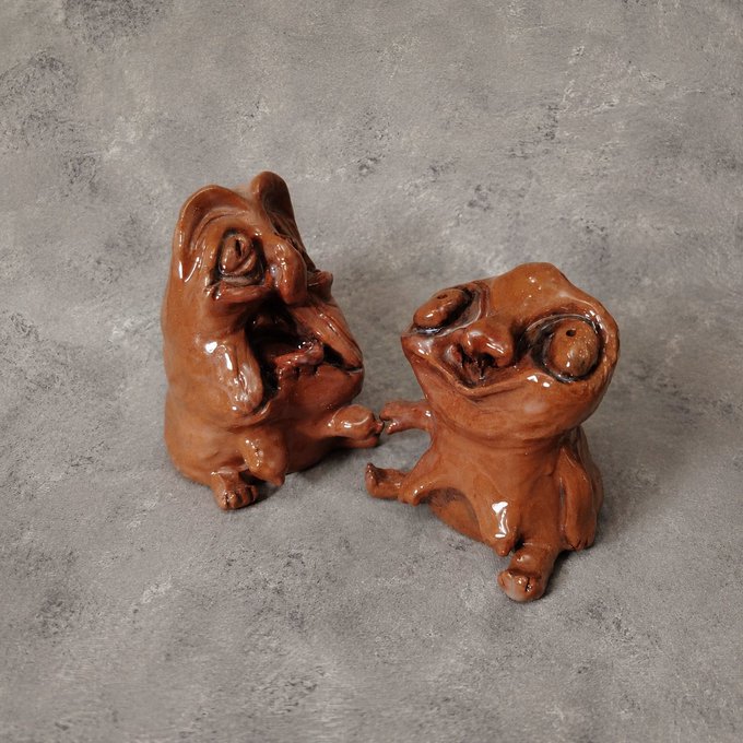 Two brown ceramic figurines depicting quirky, cartoon-like characters sit on a gray stone surface.
