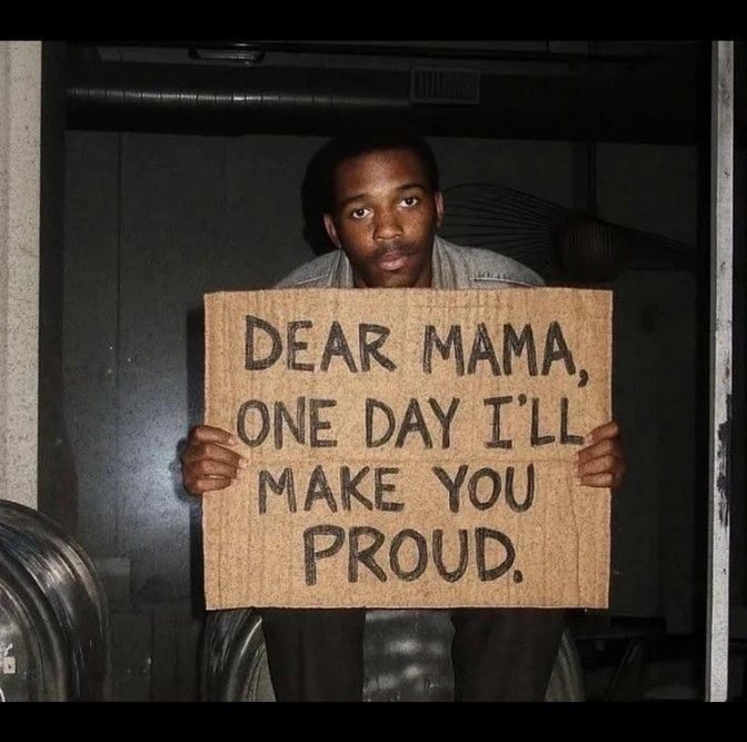 A man holds up a cardboard sign that reads 'DEAR MAMA, ONE DAY I'LL MAKE YOU PROUD.'