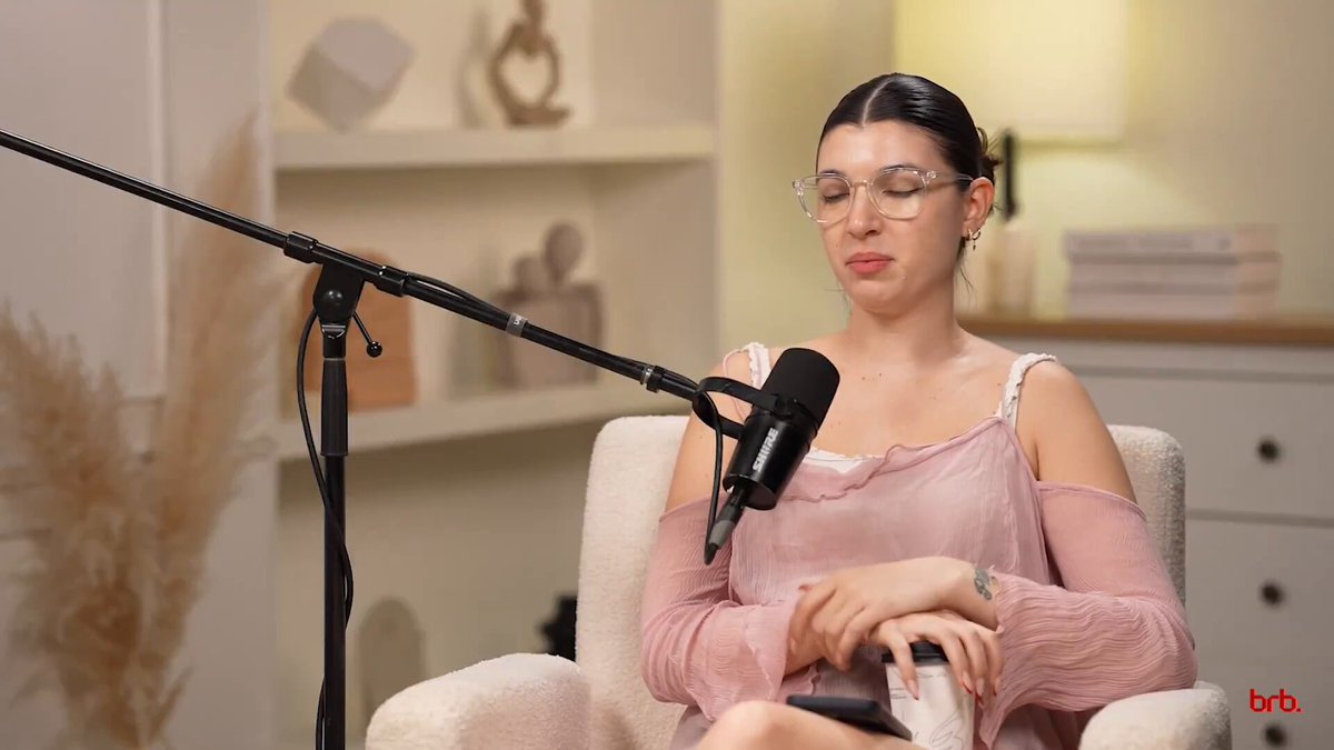 The image features a woman sitting in an armchair with her legs crossed while wearing glasses and a pink top. She appears to be talking into a microphone, possibly discussing Bitcoin or another topic related to technology. The background includes a bookshelf filled with various items such as vases and other objects.