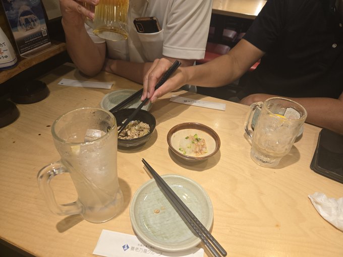 Two people sit at a wooden restaurant table with empty plates, chopsticks, mugs of beer, and a small dish of food; a hand pours more beer into a glass.