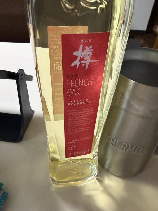 A bottle of shochu labeled "Red Queen (紅乙女) FRENCH OAK 40°" with long-term storage note sits beside a Thermos tumbler on a table. The post celebrates the user's birthday with a drink in hand while making a bullish, metaphorical reference to the rising "value" of their spirits—a lighthearted nod to the upward momentum of crypto assets like BTC and XRP.