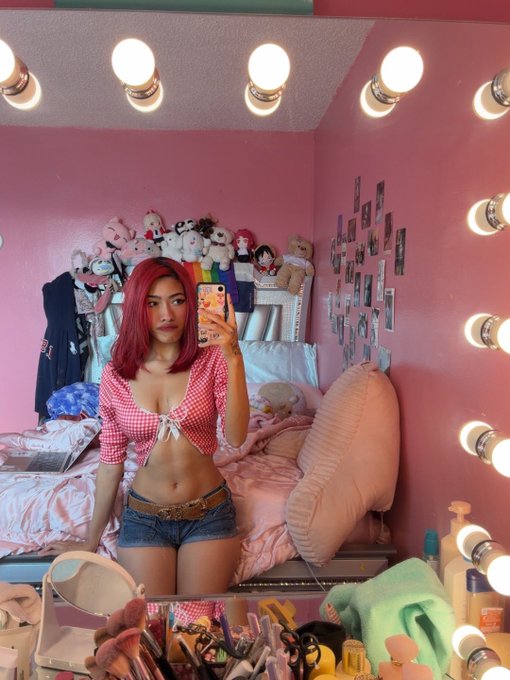 A young woman with pink hair takes a mirror selfie in her pink-themed bedroom, surrounded by vanity lights, plush toys, and polaroid photos on the wall.