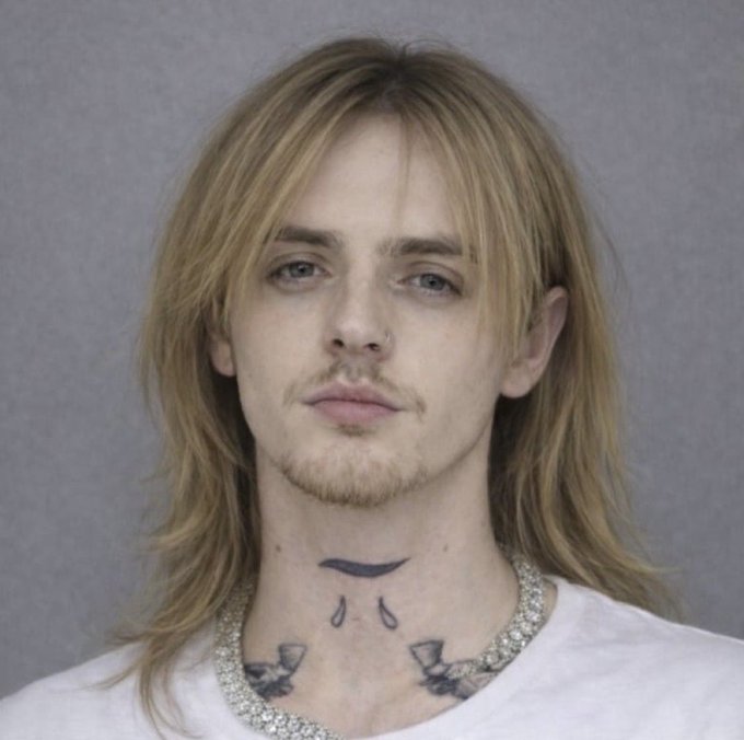 A mugshot-style portrait of a young man with long blonde hair, a nose ring, facial hair, and visible neck tattoos including teardrops and a banner.