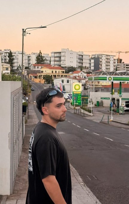 A young man with sunglasses perched on his head stands in profile on a quiet street at dawn, with a BP gas station and modern apartment buildings visible behind him. The caption "GOOD MORNING — Drop your $SOL address" fits the early morning setting of the photo, using the sunrise backdrop to set a casual, "good morning" vibe for a crypto community post asking followers to share their Solana wallet addresses.