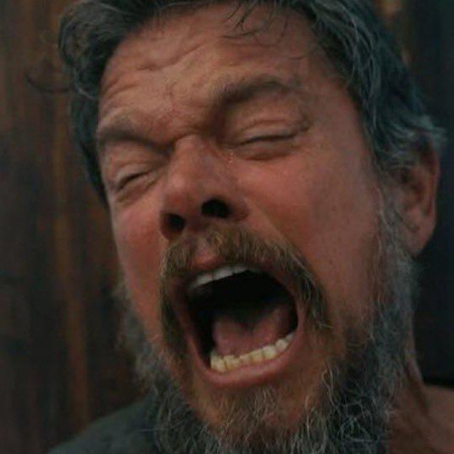 A close-up image of a bearded man with his eyes closed and mouth wide open, appearing to be screaming or crying out in intense emotion.