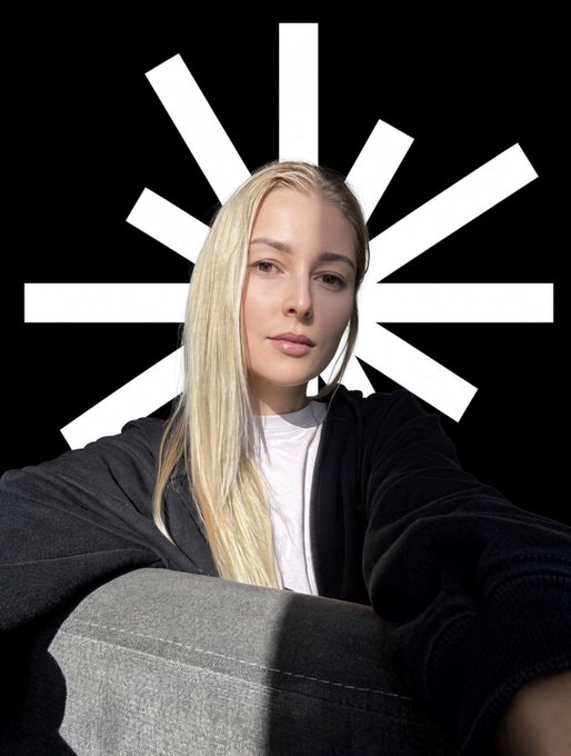 A selfie of a blonde woman wearing a dark hoodie, taken against a black background featuring a white stylized sunburst logo.