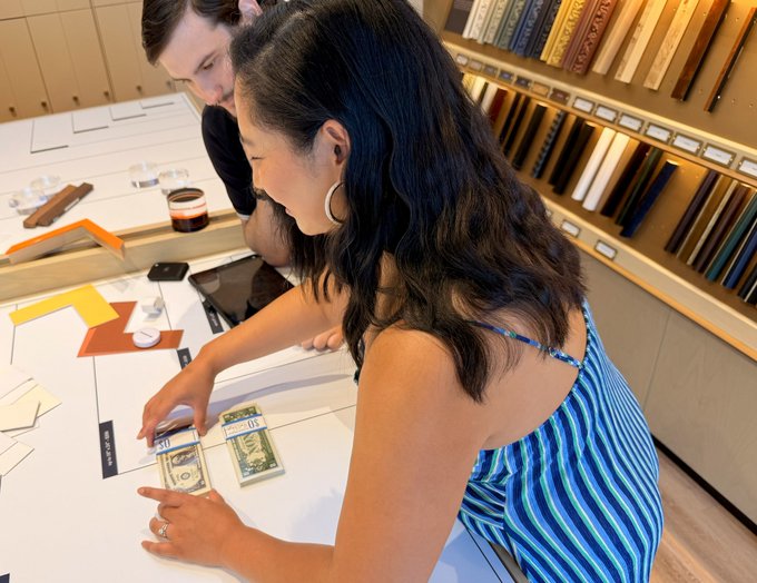 A young woman in a blue striped sleeveless top examines currency samples at a desk alongside a male colleague, with bookshelves of materials in the background.