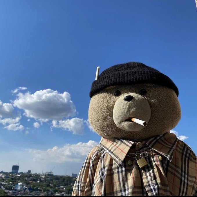 A teddy bear wearing a black beanie and plaid shirt sits outdoors under a bright blue sky, appearing to smoke a cigarette. The image serves as a metaphor for the "strongest builders" mentioned in the text, embodying a sense of calm, steady persistence despite external "noise."