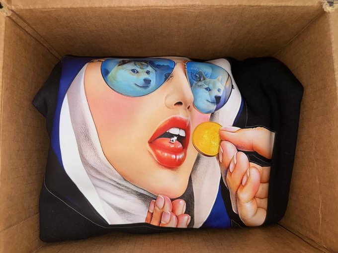 A folded T-shirt sits inside a cardboard shipping box, displaying artwork of a woman wearing sunglasses that reflect two Shiba Inu (Doge) dogs, as she holds a golden Dogecoin coin to her lips. The image visually represents the post's description of a new shirt featuring the 𝕏 logo combined with $DOGE imagery, celebrating the buyer's support for the Dogecoin community.