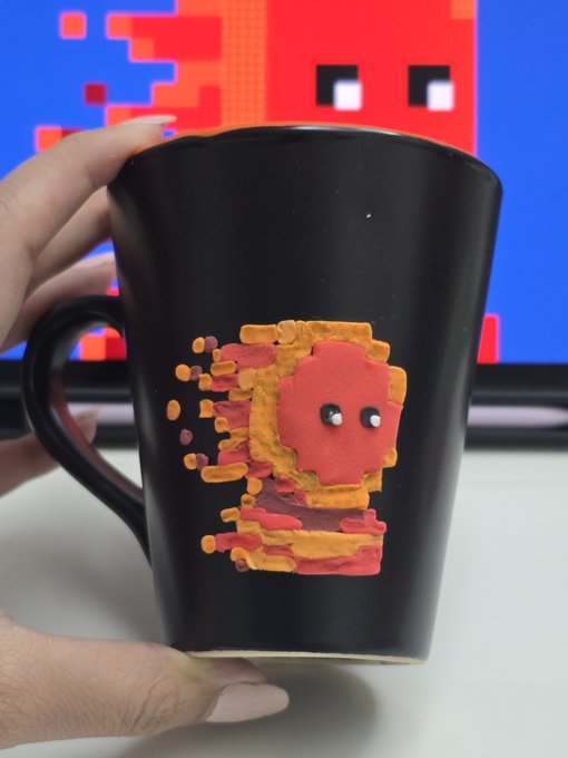 A hand holds a black coffee mug featuring a hand-painted pixel art character in red and orange, with a similar retro pixel art design displayed on a monitor in the background.