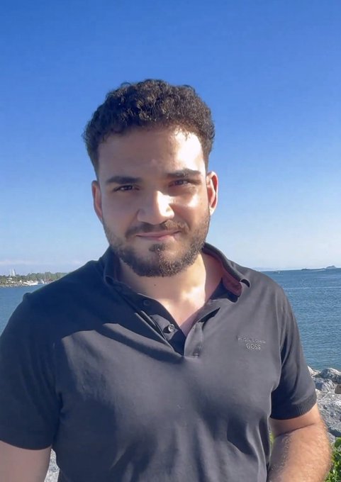 The image features a man with a beard wearing a black shirt standing near water at the beach or shoreline. The man appears to be posing for a photo while looking directly at the camera, possibly enjoying his time by the ocean. The background of the image is filled with blue sky and water, creating a serene atmosphere. There are no other people or objects in the scene, making it an interesting focal point that captures the essence of relaxation and leisure.
