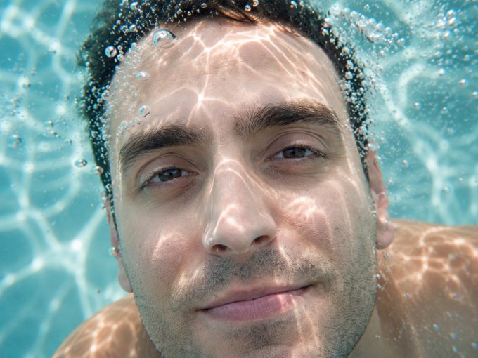 Underwater close-up portrait of a man with light eyes and stubble, surrounded by bubbles and light reflections in a turquoise pool.