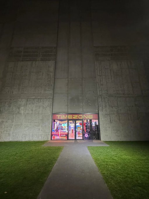 A nighttime view of a 'TIMEZONE' arcade entrance glowing with neon lights, set into a massive, bare concrete wall with a pathway and grass leading to it.