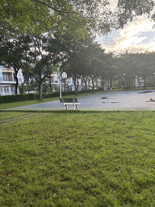 The image shows a park with a basketball court, where there are two benches placed near each other. The park also features trees surrounding the area, creating a pleasant atmosphere for outdoor activities. A building can be seen in the background, possibly indicating that this park is located within an urban setting or close to residential areas.