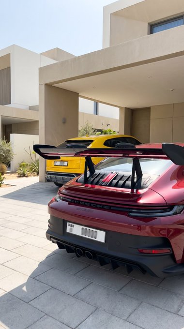 A red Porsche 911 GT3 parked in a driveway next to a yellow sports car, with a modern villa in the background.