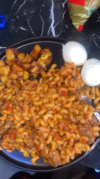 A black plate showcases a hearty meal of perfectly seasoned rotini pasta tossed with chunks of meat, alongside golden fried plantains and two peeled boiled eggs, with a colorful snack bag visible in the background. The dish serves as delicious visual proof in response to the caption's playful challenge—proving the cook's culinary skills and making it clear that calling them a bad cook is, indeed, a line not to be crossed.