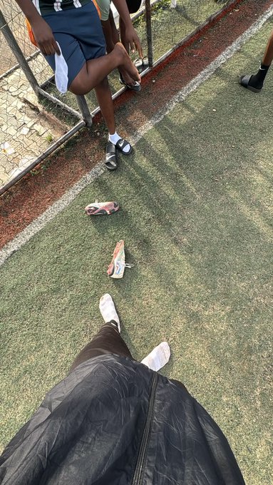 First-person view looking down at feet in white sneakers and black pants on green turf, with discarded sandals and another person's legs sitting on a railing nearby.