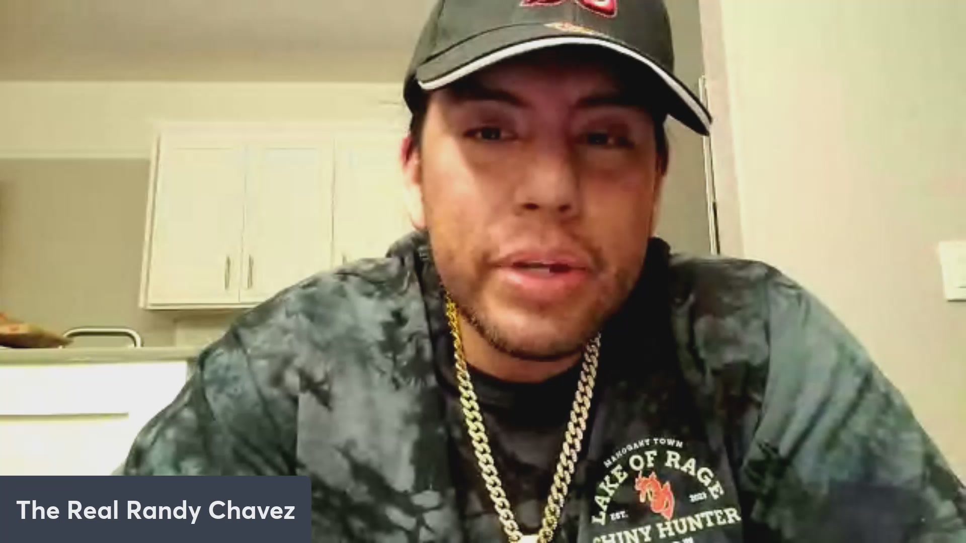 The image features a man wearing a hat, a shirt with "The Bear of Rage" written on it, and a necklace. The man appears to be talking into a microphone or camera while looking directly at the viewer. There are also some text elements in the image that read "VeVe to gross $OMI $DEFX DACAT Loki and more!".