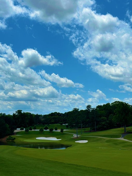 A scenic view of a golf course with a pond, sand traps, and lush green fairways bordered by trees, under a bright blue sky filled with scattered white clouds.