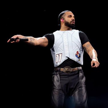 Concert photo of Drake performing on stage, wearing a white and black jersey with the number 77, holding a microphone.
