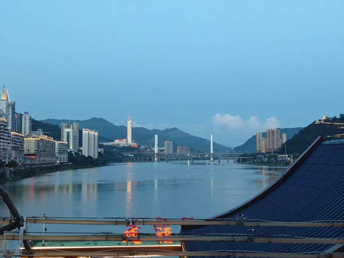 A dusk cityscape view across a river showing illuminated buildings, a bridge, and workers on a rooftop in the foreground.