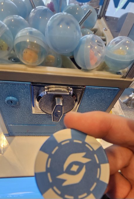 A hand holds a blue and white poker chip in front of a claw machine filled with light blue surprise capsules, with the coin slot visible on the machine's front panel.