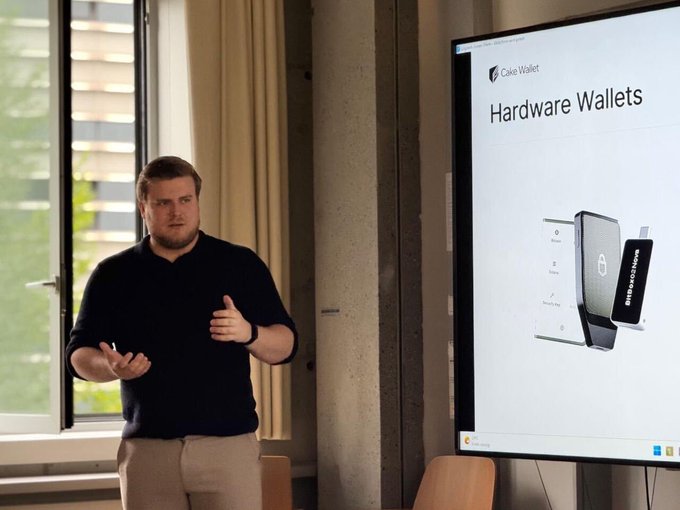 A man in a dark polo shirt stands in a classroom setting, gesturing as he presents a slide titled 'Hardware Wallets' featuring an image of a Coinbase device.