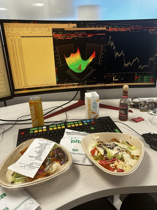 A trader's desk shows two monitors displaying a Bitcoin volatility surface and price chart, with a bowl of salad from "Fresh Pita" and a "Joyburst" drink nearby—capturing a quick lunch break while working a BTC volatility trade. The colorful 3D vol surface and candlestick chart visually reflect the "buy front BTC vol, sell back" trade mentioned in the post.