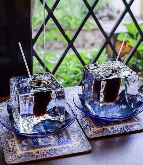 Two clear glass cups containing dark liquid sit on blue coasters by a window, with thin white straws inserted into them.