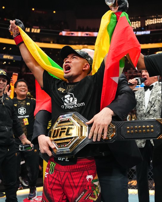 Joshua Van, draped in his flag and wearing a Crypto.com sponsored shirt, triumphantly hoists his UFC championship belt after his second title defense.