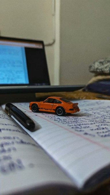 Close-up of a notebook with handwritten text, featuring a small orange Porsche toy car and a black pen, with a laptop screen visible in the background.