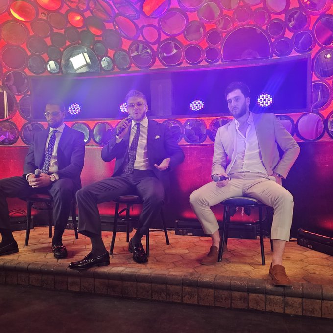 Three men in suits sitting on stools on a stage with red and purple lighting, one holding a microphone, appearing to be at a panel or event.