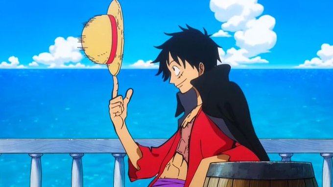 An anime character with black hair, wearing a red shirt and dark cape, balances his straw hat on his finger while sitting against a backdrop of blue ocean and sky.